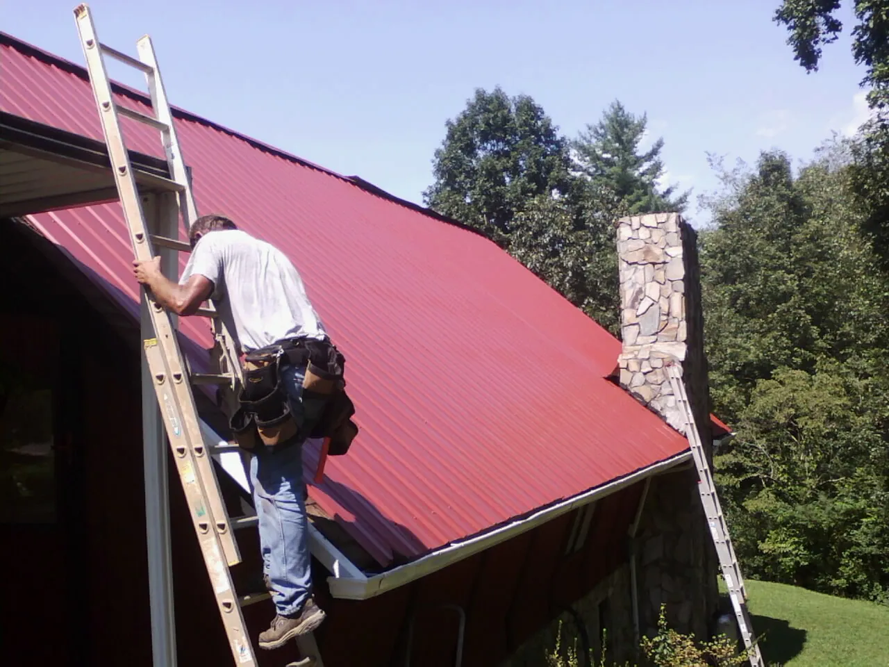 Roofer on a ladder beside a red metal roof and stone chimney — Soffit & Fascia Repair by Roofing Brothers in Prairieburg